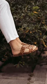 Sandalia plana confeccionada en cuero, color tostado, con tiras anchas en el empeine decoradas con tachas semiesféricas doradas y suela baja.