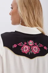 Camisa de estilo western confeccionada en viscosa color crudo. Presenta un canesú delantero y trasero en contraste color negro con bordados florales en tonos fucsia y verde, realzado con vivos en lurex dorado. Cuenta con cierre frontal mediante broches metálicos, mangas largas con puños abotonados y detalles de bolsillos decorativos en el pecho.