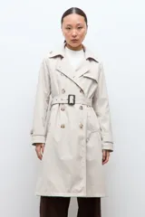 Trench coat de corte clásico en color beige, con cierre frontal de doble botonadura, solapas amplias y cinturón ajustable con hebilla. Incluye capucha desmontable y detalles de trabillas en los puños.