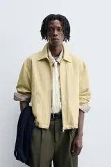 Cazadora de tejido con algodón, color amarillo pálido, con cuello solapa y cierre frontal de cremallera. Presenta bolsillos frontales de vivo y bajo ajustable con elástico en los laterales.