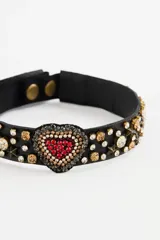 Gargantilla tipo choker de color negro, con apliques de strass y cristales en toda la banda. Presenta un dije central en forma de corazón decorado con piedras rojas y brillantes.