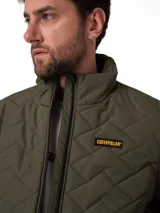 Chaqueta acolchada de color verde oliva con diseño de costuras en zigzag. Presenta cierre frontal completo, cuello alto y parche con el logo de la marca en el pecho.