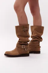 Bota de caña alta color marrón chocolate, confeccionada en gamuza. Presenta dos correas desmontables con hebillas metálicas plateadas.