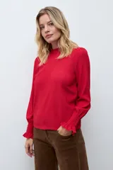Blusa de color rojo con cuello alto y detalle de volados, mangas largas con puños elásticos en punto smock y textura sutil en la tela.