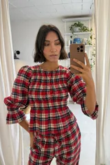 Blusa de viyela con estampado de cuadros escoceses en tonos rojo, negro y beige. Presenta un diseño con frunces en el pecho, mangas tres cuartos abullonadas con puños elásticos y un corte ajustado en la cintura que se abre en un pequeño volante.