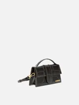 Bolso Le Grand Bambino de Jacquemus confeccionado en cuero de ternera con estampado efecto cocodrilo en color marrón oscuro. Presenta una silueta estructurada, solapa frontal con cierre magnético y tira vertical, asa superior rígida y correa ajustable y desmontable. Incluye detalles metálicos dorados con el logotipo de la marca en el frente.