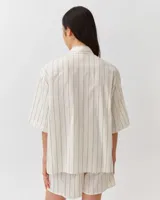 Camisa de corte recto y diseño depurado, color blanco con finas rayas verticales oscuras. Presenta cuello camisero clásico, cierre frontal de botones ocultos y mangas cortas de silueta relajada.