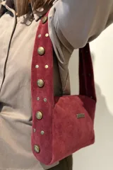 Cartera de hombro confeccionada en gamuza color bordó, con correa ancha decorada con tachas metálicas circulares de distintos tamaños. Presenta un diseño estructurado con cierre superior y placa metálica pequeña con logo en el frente.