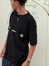 Remera negra de corte oversize con cuello redondo y manga corta. Presenta tres estampados blancos en el frente: un símbolo curvo a la izquierda, la palabra "OSTEN" en el centro y un símbolo abstracto a la derecha.