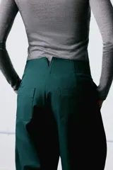 Pantalón de tiro alto confeccionado en mezcla de algodón, con diseño de volumen y costuras torcidas. Presenta bolsillos laterales y cierre frontal con cremallera, botón interior y ganchos metálicos ocultos.