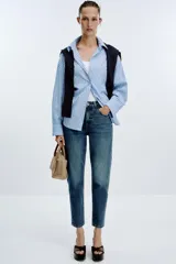 Jeans de mujer, tiro alto, corte Mom Fit con pierna slim y largo tobillero. Denim azul medio con efecto desgastado y lavado. Presenta cinco bolsillos y cierre frontal con cremallera y botón.