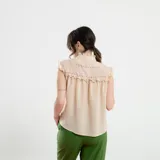 Camisa sin mangas color beige, con cuello alto con volados, detalles de encaje calado en los hombros y parte superior del pecho, y cierre frontal con botones.