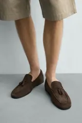 Mocasines de vestir para hombre confeccionados en piel con acabado en gamuza de color marrón, con detalle de borlas en el empeine y suela plana con ligero tacón.