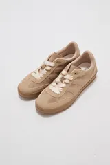 Zapatillas urbanas de corte bajo en tonos beige y tostado, combinando paneles de cuerina y gamuza sintética. Cuentan con cordones anchos tipo cinta de raso en color crudo, pespuntes decorativos y suela de goma al tono.