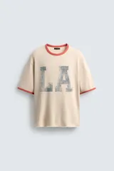 Camiseta de punto tejida color beige claro, corte boxy fit, con cuello redondo y mangas cortas. Presenta estampado desgastado con las letras "LA" en el frente y ribetes en color terracota en cuello y mangas. El bajo es irregular.