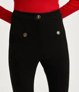 Pantalón flare negro de punto roma, con cintura alta, bolsillos delanteros con botones metálicos estilo escudo y cierre por cremallera y botón.