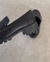 Mocasines negros de estilo clásico con textura de cuero grabado tipo cocodrilo. Presentan una banda frontal característica y suela plana con borde definido.