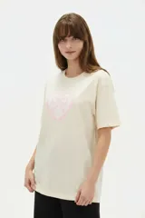 Remera de algodón color beige con estampado frontal de un corazón rosado que contiene un moño. Tiene cuello redondo y mangas cortas.