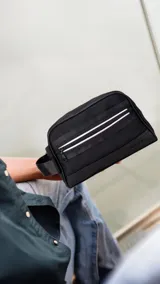 Neceser negro rectangular con cierre superior y bolsillo frontal con cremallera. Tiene asa lateral y dos bandas horizontales blancas en el frente.