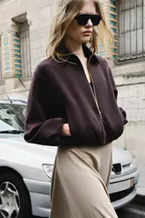 Chaqueta de corte corto y diseño abullonado en color marrón oscuro, con cuello solapa, mangas largas con puños elásticos y cierre frontal con cremallera metálica. Cuenta con bolsillos delanteros de doble vivo.
