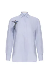 Camisa de lino celeste con cuello camisero, manga larga y cierre frontal oculto con botones. Incluye un broche desmontable bordado con forma de colibrí y plumas en el pecho.