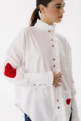 Camisa blanca de algodón con cuello alto con volados, cierre frontal con botones, mangas largas con puños abotonados, bolsillo en el pecho con corazón bordado y parches de crochet en los codos.