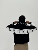 Canguro con capucha de corte boxy, color negro con una franja horizontal blanca que recorre la espalda y las mangas, presentando la palabra 'BROOKLYN' estampada en negro sobre la franja. La capucha también incluye detalles estampados.