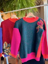 Sweater de punto con diseño color block, presentando un frente en tono verde oscuro melange con un bordado floral en el hombro, y mangas en color fucsia vibrante. Posee cuello redondo y puños acanalados.