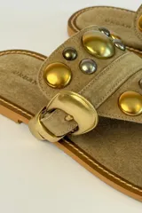 Sandalia plana de gamuza marrón, estilo ojota, con apliques metálicos dorados y hebilla decorativa.