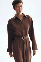 Vestido midi marrón de estilo camisero, con cuello, cierre frontal de botones, hombros caídos y mangas largas. Incluye un cinturón de tela con hebilla metálica de anillas en D.