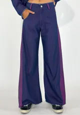 Pantalón de jean azul con corte ancho y una franja vertical de color violeta en los laterales. El ruedo está desflecado.