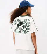 Remera boxy de algodón color blanco, con cuello redondo y ribete verde oscuro, manga corta con líneas decorativas en los hombros, y estampado frontal deportivo de Mickey Mouse con tipografía.