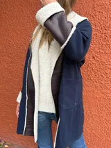 Saco de paño azul marino con solapas amplias y puños en corderito sintético color crema. Presenta detalles en cuero sintético marrón en la parte frontal y en las mangas, con un diseño abierto y corte recto.