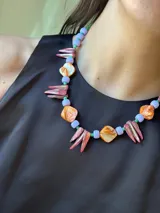 Collar corto con cuentas de nácar teñido en tonos anaranjados y rosados, piedras naturales, cuentas de acrílico lilas y mostacillas verdes. Se cierra con cadena de acero quirúrgico y ganchito para regular el largo.