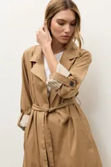 Trench de lyocell color beige con solapas amplias, cierre cruzado con botones, bolsillos laterales y mangas largas con presillas en los puños que permiten ajustarlas.