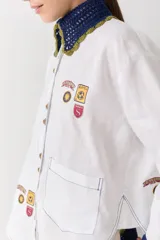 Camisa blanca de corte holgado con cuello y puños tejidos en crochet azul con borde verde. Presenta parches bordados decorativos en el frente y las mangas, cierre frontal con botones y un bolsillo de parche en el pecho.
