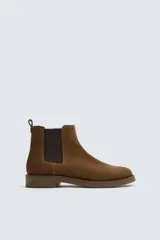 Bota estilo Chelsea de hombre, color marrón, fabricada en piel con acabado serraje (gamuza). Presenta elásticos laterales oscuros y suela de goma a tono con tacón bajo.