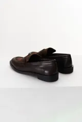 Mocasín color marrón claro, con capellada de cuero liso y aplique de gamuza en marrón oscuro. Tiene punta redondeada y suela plana de goma negra.