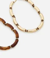 Collar de doble capa con diseño de tubitos curvos. Una de las cadenas presenta tubitos con acabado dorado brillante, mientras que la otra cuenta con tubitos de resina translúcida en tono ámbar, ambos intercalados con pequeñas cuentas metálicas. Posee un cierre de mosquetón y está confeccionado con materiales reciclados.