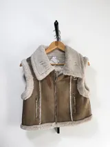 Chaleco estilo gamulán con interior y bordes de piel sintética. Presenta cierre frontal metálico, bolsillos laterales con cierre y detalles de costuras visibles.