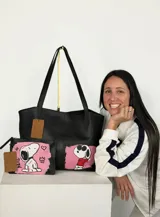 Tote bag de cuero negro con diseño intervenido a mano por la artista GGIL, presentando ilustraciones de Snoopy sobre fondos rosados. Incluye un sobre a juego con el mismo diseño artístico.
