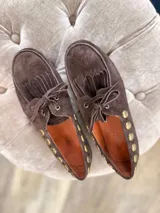 Náuticos de gamuza color verde oliva, con diseño de flecos en la parte frontal, cordones y detalle de tachas metálicas doradas en los laterales. Calzado artesanal con interior forrado.