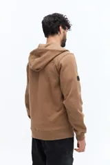 Campera de felpa color camel con cierre frontal, capucha ajustable con cordones negros y bolsillos laterales con cierre negro. Presenta un estampado pequeño en el pecho con texto en color oscuro.