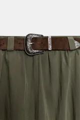 Falda midi tipo capa de tiro medio, confeccionada en mezcla de algodón color verde militar. Cuenta con cintura con trabillas, bolsillos de vivo en los laterales, cierre lateral con cremallera oculta y aplicación de cinturón marrón de estilo western con hebilla metálica grabada.