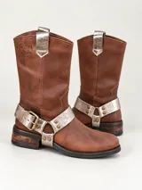 Bota de caña alta de cuero marrón, con taco bajo y detalles plateados en relieve texturizado. Cuenta con una hebilla metálica grande y tiras desmontables con tachas decorativas alrededor del tobillo, además de una presilla superior en la parte trasera.