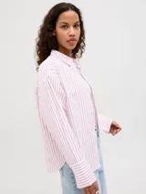 Camisa de popelín de manga larga con cuello clásico y cierre frontal de botones. Presenta un diseño de rayas verticales finas en color rosado sobre fondo blanco, con un corte relajado y puños anchos.