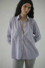 Camisa de rayas verticales rosadas y celestes, con cuello clásico, botonadura frontal y doble bolsillo delantero. Tiene un calce relajado y terminación curva en el ruedo.