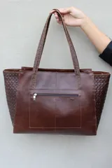Bolso tipo shopper de gran tamaño confeccionado en cuero vacuno color marrón oscuro. Presenta laterales con textura grabada tipo cocodrilo y detalles de costuras decorativas con tientos de cuero en los paneles frontales y traseros. Incluye doble asa de hombro, cierre superior y placa metálica decorativa en el frente.