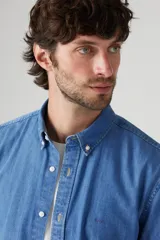 Camisa de manga larga de denim azul claro, con cuello abotonado (button-down) y botones frontales blancos.