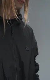 Campera rompevientos negra de calce oversize, confeccionada en microfibra técnica ligera con acabado semi-mate. Presenta capucha integrada, cierre frontal técnico y bolsillos tipo ojal. Destaca por su sistema de frunces elásticos regulables mediante tancas a lo largo de las mangas y el ruedo, permitiendo ajustar el volumen de la prenda.